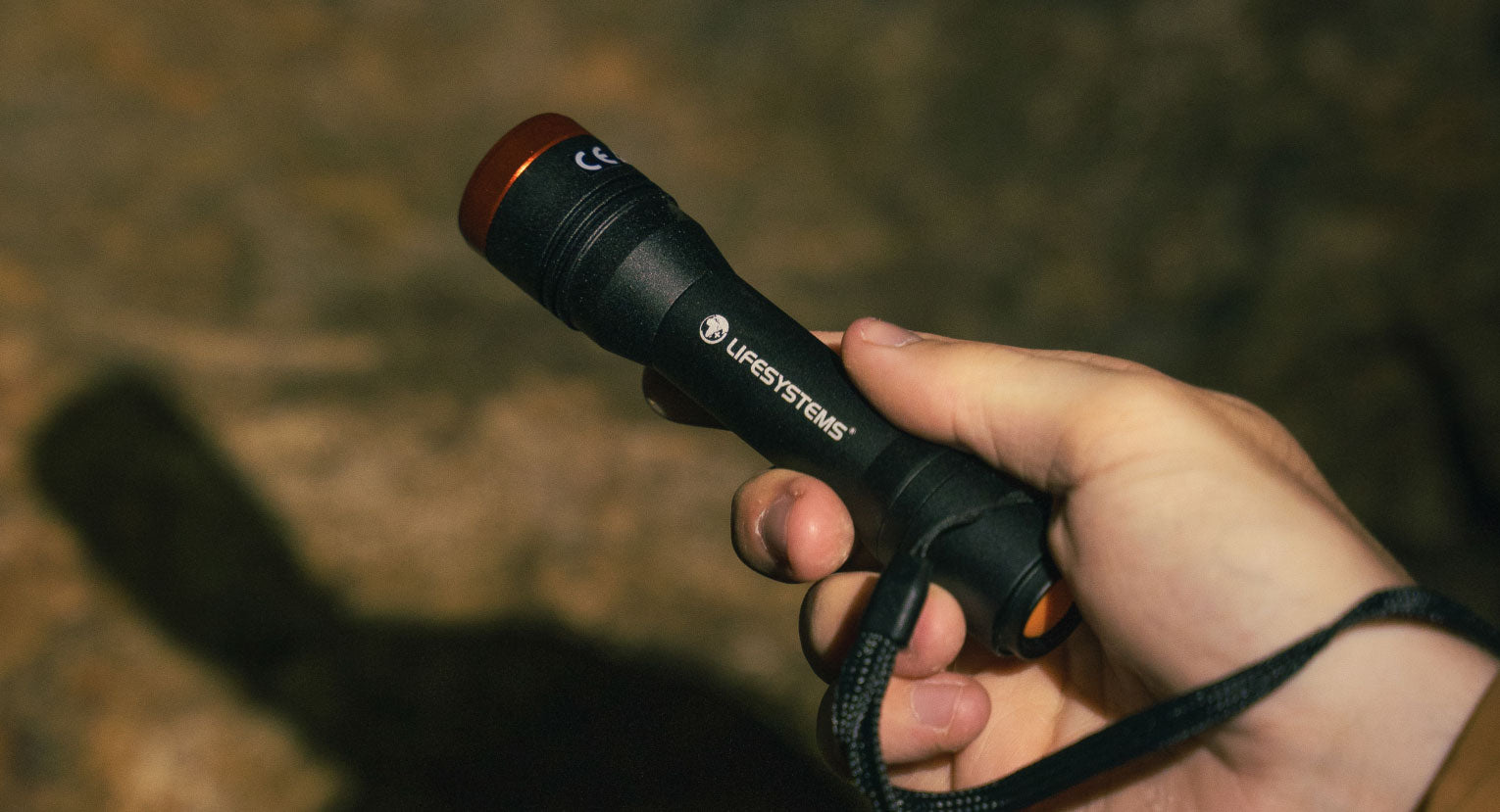 Person holding a Lifesystems Intensity 480 hand torch in a dimly lit outdoor setting, casting a visible beam and shadow on the ground.